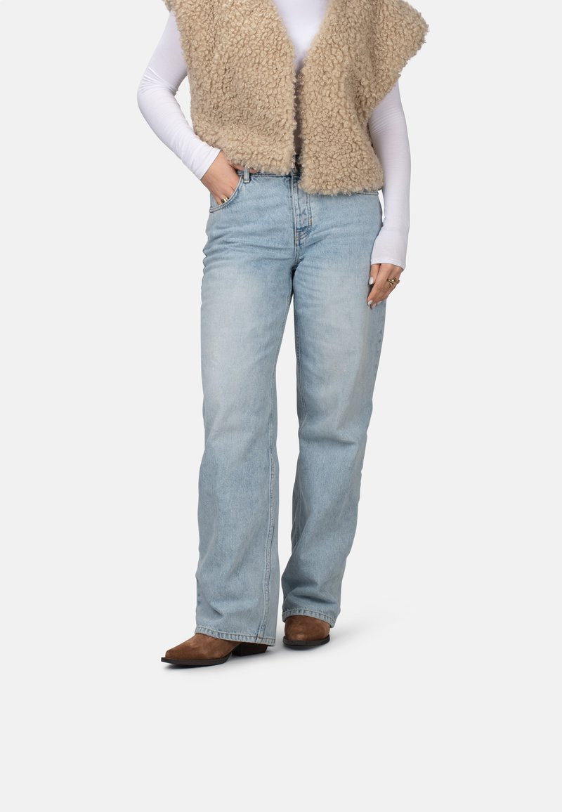 Chaleco de piel sintética beige sobre una camisa blanca de manga larga, combinado con vaqueros sueltos de color azul claro y botas de tobillo marrón con tacón.