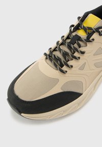 Baskets de sport beiges avec une tige en maille texturée, un capuchon en caoutchouc noir à l'avant, et des accents noirs et jaunes sur la languette. Présentent des lacets à motifs.