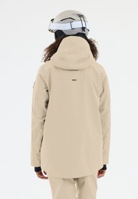 Beige vattent�ät jacka med hög krage och justerbar huva, med en logotag på baksidan och långa ärmar. Slät textur.