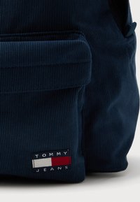 Mørkeblå kordfløjl rygsæk med en frontlomme og et broderet logo-tag med røde, hvide og blå striber. Lynlås lukning i toppen.