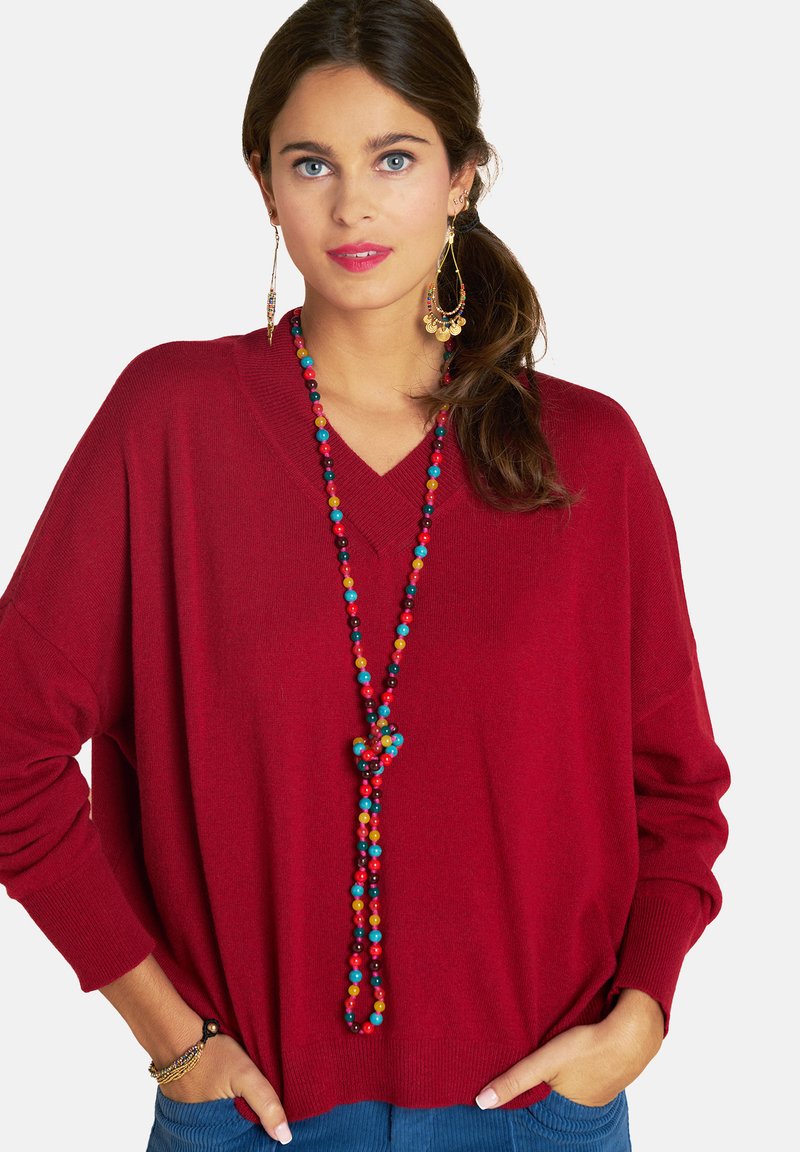 Roter Strickpullover mit V-Ausschnitt, kombiniert mit einer mehrfarbigen Perlenkette und großen Ohrringen. Glatte Textur, lockere Passform, lässiger Stil.