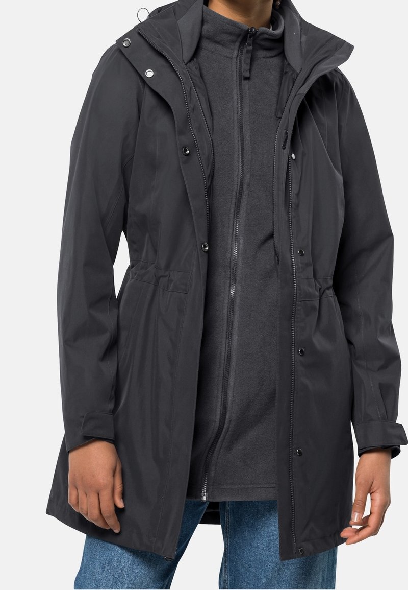 Musta, vedenpitävä takki, jossa on nyörillinen huppu, napit ja joustava vyötärö. Päällä harmaa vetoketjullinen fleece ja siniset farkut.
