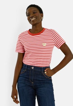 Mujer sonriente con camiseta a rayas rojas y blancas con detalle de corazón dorado, vaqueros oscuros de talle alto y joyas doradas, mano en la cadera.