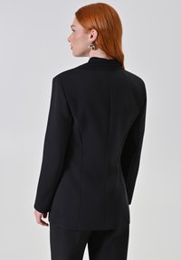 Blazer nero su misura, forma aderente, spalle strutturate, texture liscia, con colletto alto e dettagli di cucitura sottili sul retro.