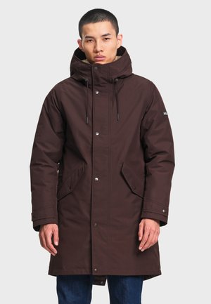 Man draagt een donkerbruine winterjas met capuchon en klepzakken met knopen, staand tegen een eenvoudige lichte achtergrond.