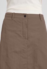 Jupe marron avec une texture lisse, poche avant, passants de ceinture et fermeture à bouton. Le tissu présente un aspect légèrement froissé.