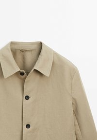 Giacca beige chiaro con colletto, caratterizzata da tre bottoni neri e un tessuto liscio e testurizzato. Cucitura laterale visibile, senza motivi o accenti.
