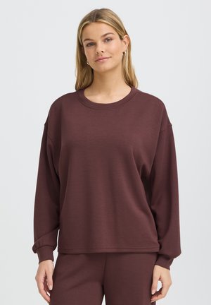 Jeune femme aux cheveux blonds et raides portant un sweat-shirt ample à manches longues marron foncé et un pantalon assorti sur fond blanc.