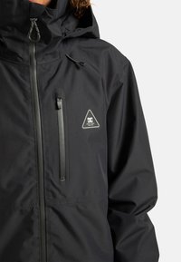 Czarna wodoodporna kurtka z zamkiem błyskawicznym na kieszeni piersiowej, regulowanym kapturze i trójkątną łatą logo po lewej stronie. Gładka struktura.