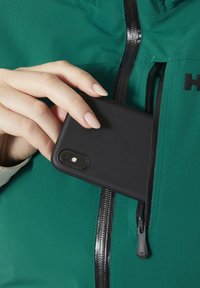 Černý obal na smartphone, který je držen nad zipovou kapsou na zelené bundě. Kapsa má černý zip a hladký povrch.