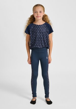 Junges Mädchen mit lockigem Haar steht und trägt ein marineblau gemustertes T-Shirt sowie passende Leggings und schwarze Ballettschuhe vor grauem Hintergrund.