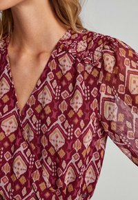 Roter Stoff mit geometrischen Mustern in Rosa, Gelb und Weiß. Verfügt über einen V-Ausschnitt und geraffte Details an den Schultern.