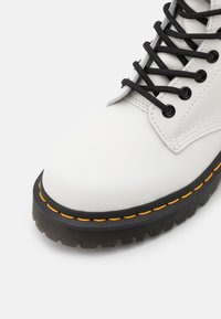 Botte en cuir blanc avec semelle épaisse noire et surpiqûres jaunes, dotée de lacets noirs passés dans des œillets métalliques.