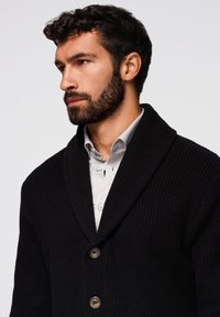 Czarny sweter dzianinowy z kołnierzem szalowym, z dwoma guzikami, o teksturowanej tkaninie, noszony na jasnoszarej koszuli.