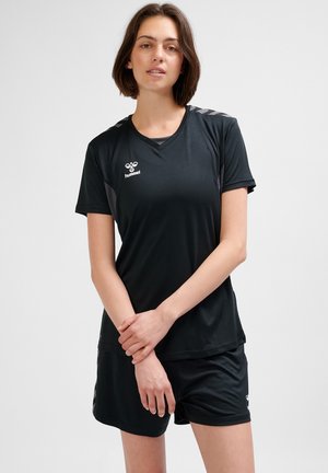 Mujer joven con camisa deportiva negra de manga corta y pantalones cortos, de pie frente a un fondo claro y liso con las manos suavemente entrelazadas.