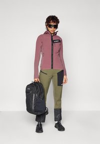 Veste légère, à manches longues, couleur mauve avec fermeture éclair. Un pantalon vert avec des accents noirs, portant un sac à dos noir. Lunettes de soleil sombres portées.