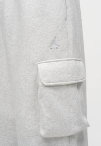Tissu gris clair avec une poche à rabat et un logo Jumpman Air brodé au-dessus de la poche sur une manche de vêtement.