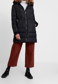 Abrigo negro acolchado con cremallera frontal, cintura ajustable con cordón elástico y capucha de piel sintética. Combinado con pantalones anchos marrones y botas negras con cordones.