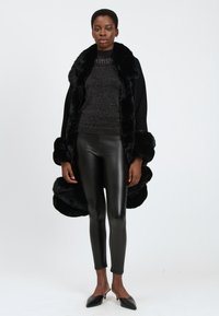Abrigo de pelo sintético negro con un amplio cuello y puños, llevado sobre un suéter negro brillante, combinado con leggins de cuero negros y tacones de punta.