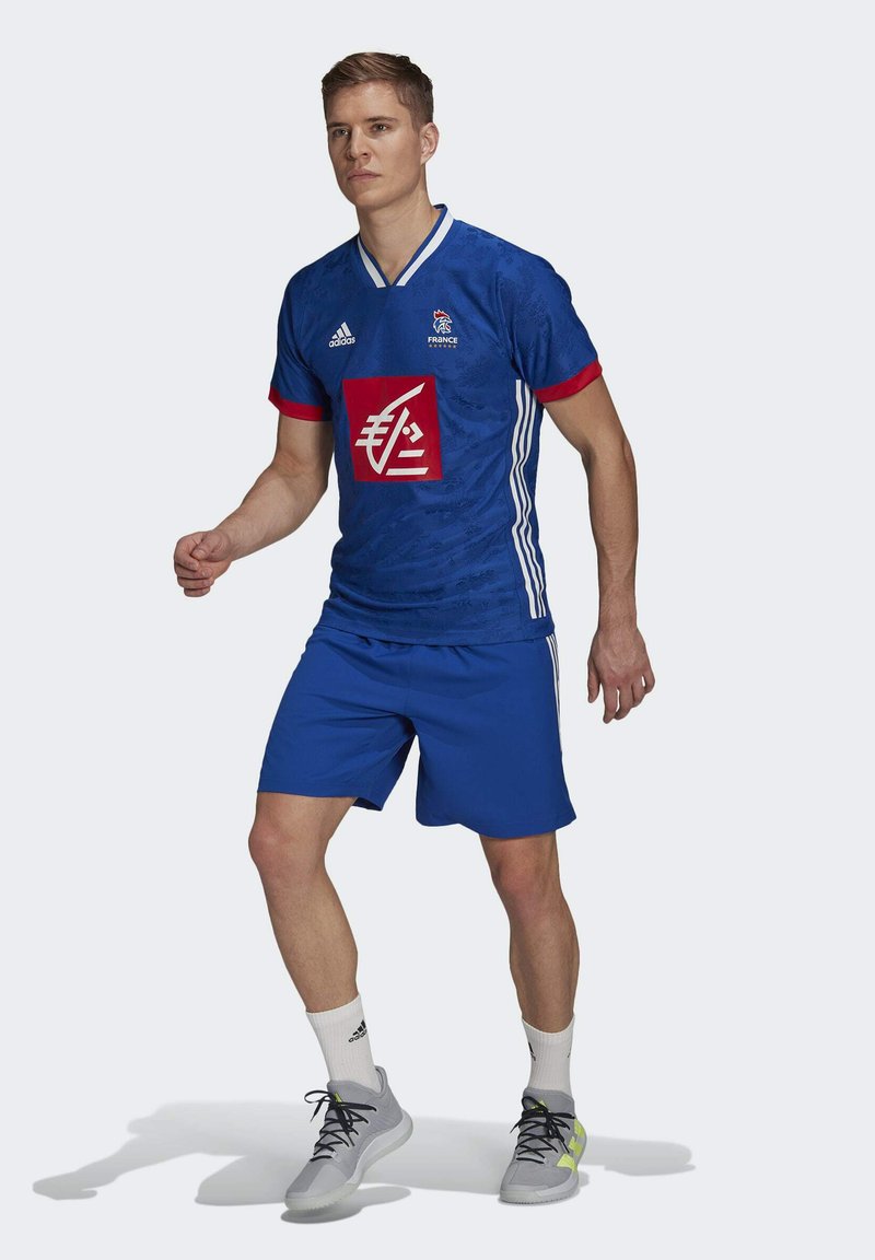 Blauw voetbalshirt met rode accenten en witte strepen, voorzien van een groot rood vierkant logo. Gecombineerd met blauwe shorts en grijze sportschoenen.