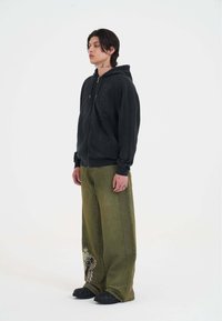 Schwarze Zip-Up Kapuzenjacke mit strukturiertem Stoff und großem Logo-Druck. Lockere olivegrüne Hose mit gesticktem Adlerdesign. Schwarze Schuhe.