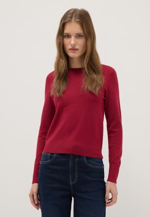 Giovane donna con lunghi capelli castano chiaro che indossa un maglione rosso semplice a maniche lunghe e jeans blu scuro su uno sfondo neutro.