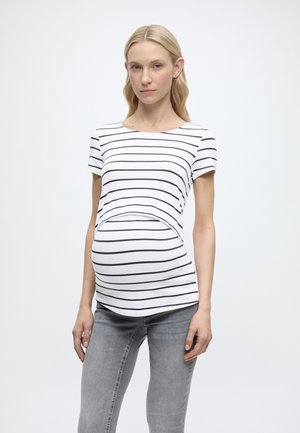 Terhes nő fehér-fekete csíkos rövid ujjú felsőben és szürke farmerben, sima háttér előtt áll.