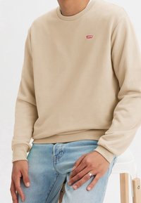 Beige crewneck sweatshirt i mjukt tyg, med en liten röd Levi's-logotyp. Kombinerad med ljusblå denimjeans.