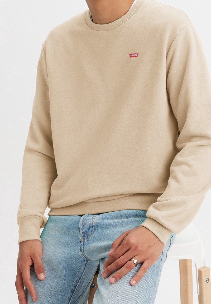 Beige crewneck sweatshirt i mjukt tyg, med en liten röd Levi's-logotyp. Kombinerad med ljusblå denimjeans.