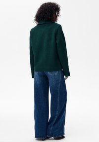 Kobieta z kręconymi włosami, ubrana w ciemnozieloną kurtkę, niebieskie szerokie dżinsy, czarne botki i okulary przeciwsłoneczne, stojąca tyłem na białym tle.