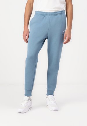 Person iført lyseblå sweatpants med elastisk talje og ankler, kombineret med hvide sneakers og en hvid skjorte mod en ensfarvet baggrund.