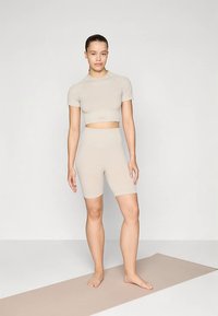 Beigen sävyinen, yhteensopiva treeniasusetti, joka sisältää lyhythihaisen, lyhennetyn topin ja korkeavyötäröiset pyöräilyshortsit. Valmistettu pehmeästä, joustavasta kankaasta.