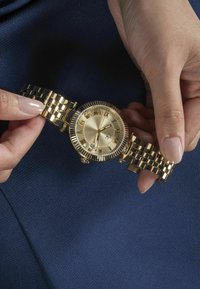 Gul armbandsur med en strukturerad rund urtavla, romerska siffermarkeringar och ett metall länkbandsarmband. Utrustad med en rund krona och dekorativa detaljer.