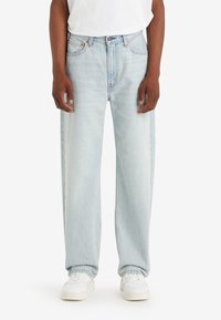 Uomo che indossa jeans a gamba dritta blu chiaro e sneakers bianche, in piedi contro uno sfondo bianco semplice.