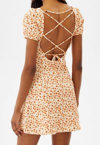 Robe mini fleurie crème à fleurs orange, avec manches courtes bouffantes et dos croisé à nouer. Tissu texturé et léger.