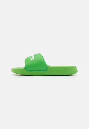 Sandale en caoutchouc vert avec une large sangle portant un texte blanc. Texture lisse avec une semelle plate et de petits détails de griffes circulaires.