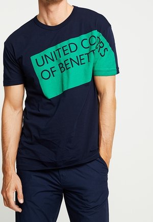 Mężczyzna ubrany w granatowy t-shirt z zielonym ukośnym pasem i czarnym napisem "UNITED COLORS OF BENETTON", w zestawieniu z granatowymi spodniami.