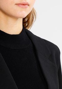 Jersey negro de cuello alto acanalado combinado con un blazer negro entallado, que muestra una textura suave y un diseño estructurado.