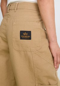 Khaki cargo pants in sturdy fabric, featuring a back pocket with a black label that reads "ALPHA INDUSTRIES EST. OCTOBER 1959".