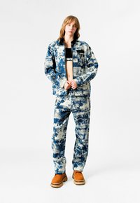 Conjunto de chaqueta y pantalones de mezclilla en un patrón abstracto azul y crema; la chaqueta cuenta con un cierre de cremallera y un bolsillo en el pecho; se combina con botas naranjas.