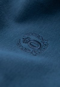 Close-up af blåt stof med et diskret broderet rundt logo med en krans og en oval form i midten.