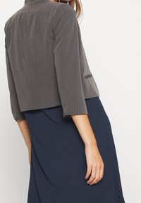 Harmaa lyhyt takki kolmannelta vartalolta, muotoiltu malli, yksi tasku, puettuna tummansinisen mekon päälle. Sileä pinta, kevyt materiaali.