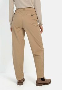 Beige, weit geschnittene Hose mit glatter Textur, ausgestattet mit einer Gesäßtasche und einem dekorativen Knopf. Stilisiert mit braunen Stiefeletten.