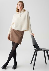 Cremfarbener, gerippter Strickpullover mit Halsreißverschluss, kombiniert mit einem braunen Wildleder-Minirock, schwarzen Strumpfhosen und schwarzen Mokassins. Schwarzer Stuhl im Hintergrund.