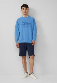 Man in een lichtblauwe sweatshirt met de tekst "S.Oliver", marineblauwe knielange shorts en witte sneakers tegen een effen grijze muur.