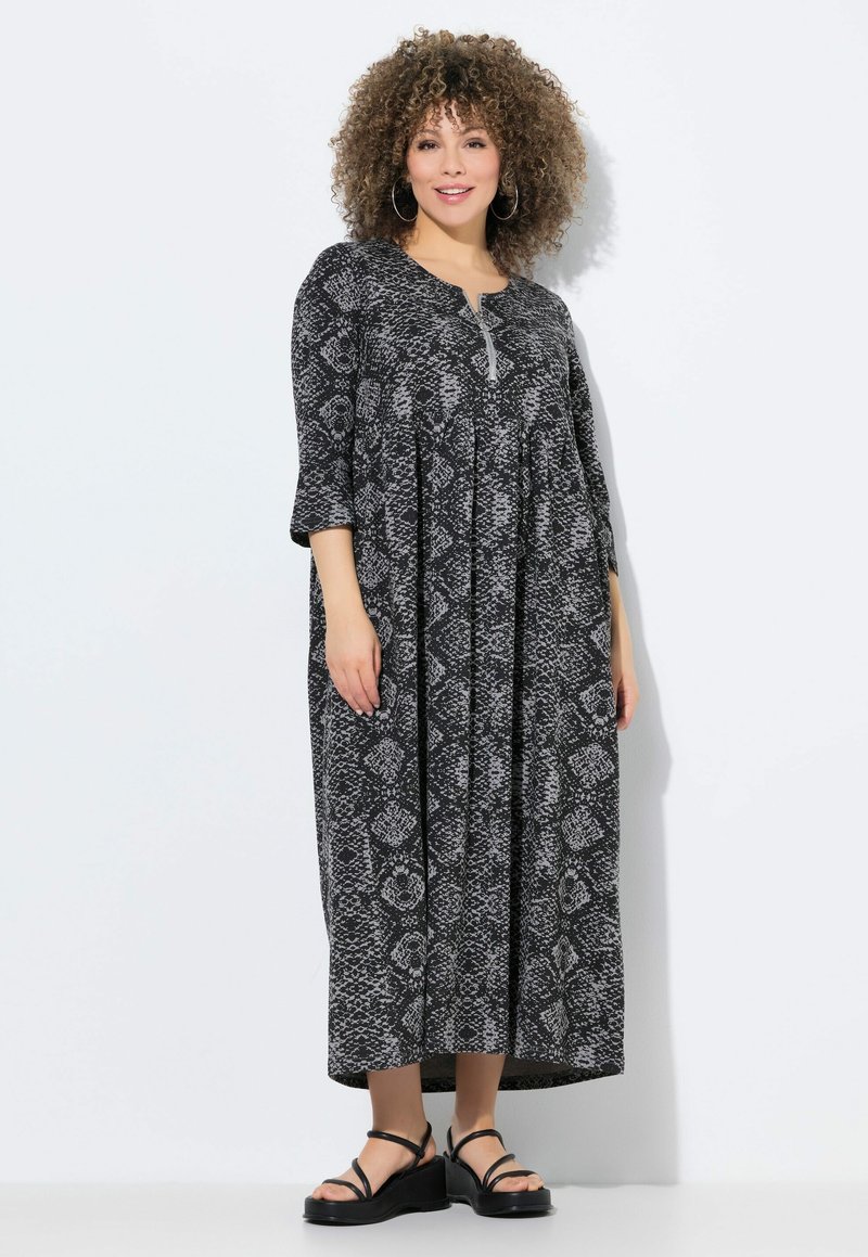 Schwarz-weißer Schlangenprint-Kleid mit Dreiviertelärmeln, rundem Ausschnitt und einem Knopfdetail an der Brust. Modell trägt schwarze Sandalen.