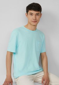 Lichtblauw T-shirt met korte mouwen van een zachte stof, met een ronde halslijn en een klein borstzakje. Model zit tegen een neutrale achtergrond.