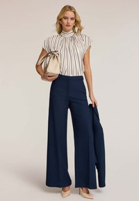 Blusa a strisce crema con colletto alto, abbinata a pantaloni larghi blu navy. Tiene una borsa beige e indossa tacchi nudi.
