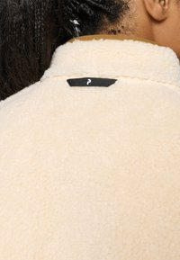 Gros plan sur une personne portant une veste en polaire de couleur crème avec une étiquette en tissu noir à l'arrière du col, montrant des cheveux tressés et une oreille.