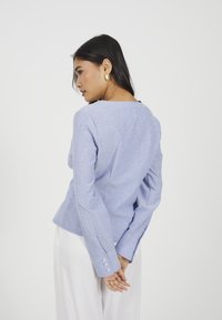 Donna con lunghi capelli scuri che indossa orecchini d'oro, una maglia blu chiaro a maniche lunghe con bottoni ai polsini e pantaloni bianchi, con le mani unite dietro la schiena.